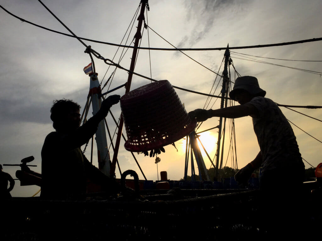 Two people silhouetted against the background of a ship or shipyard are emptying a large basket over their heads of fish.